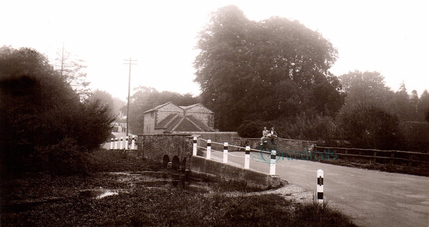 Old photos of  girls sitting on  river Lavant bridge wall with little boy between them, bicycle leaning on wall, bus in distance, Chichester - further image below